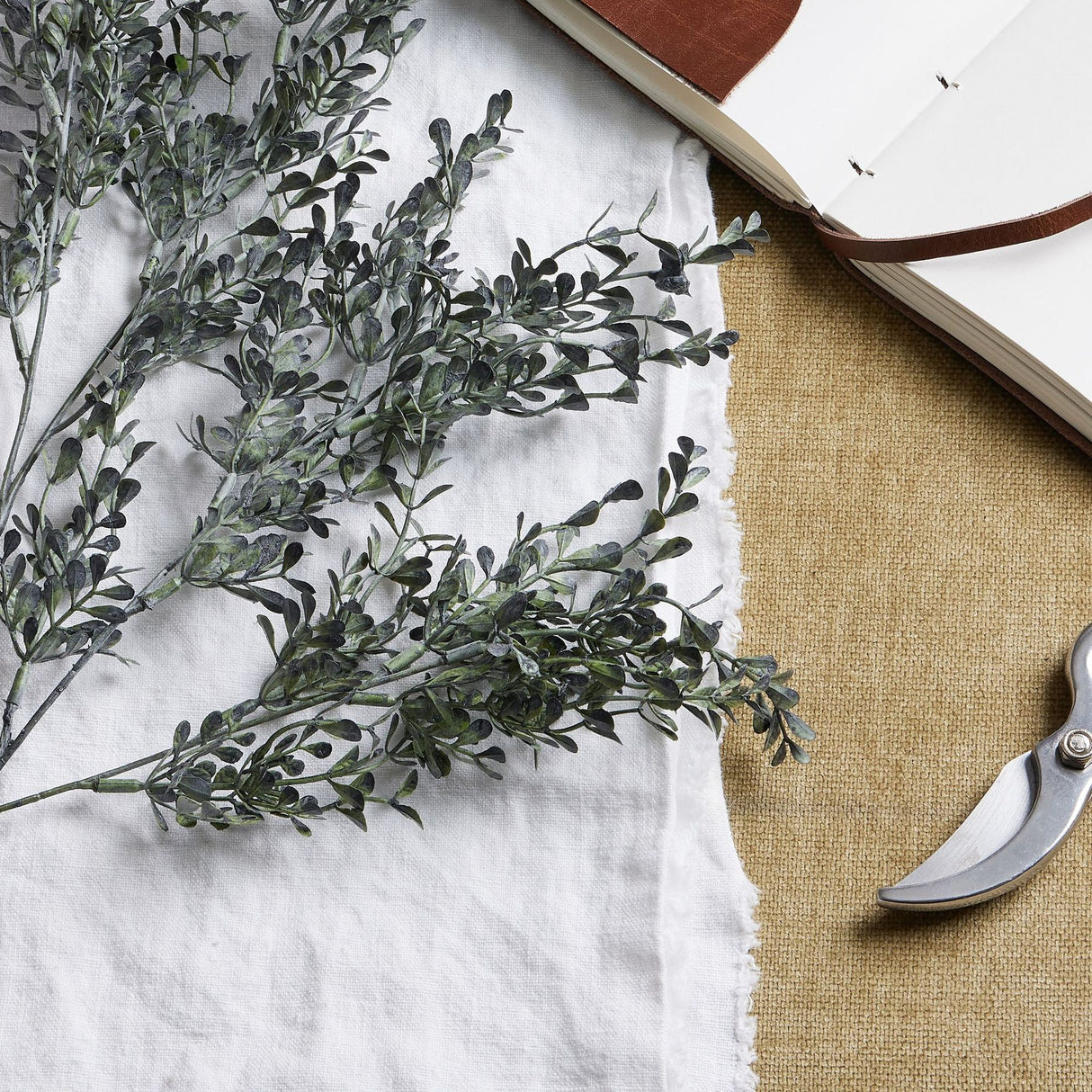 Hill Interiors Blackened Grass Spray Stem with dark feathered foliage laid on white linen beside an open leather-bound journal and metal secateurs.
