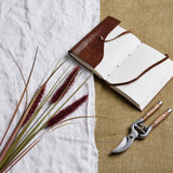 Ruby Triple Grass Stem by Hill Interiors, an 80cm faux dried grass with red plumes, styled on white linen beside a leather journal and wooden secateurs.
