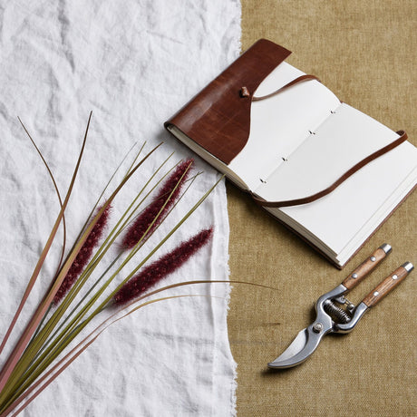 Ruby Triple Grass Stem by Hill Interiors, an 80cm faux dried grass with red plumes, styled on white linen beside a leather journal and wooden secateurs.