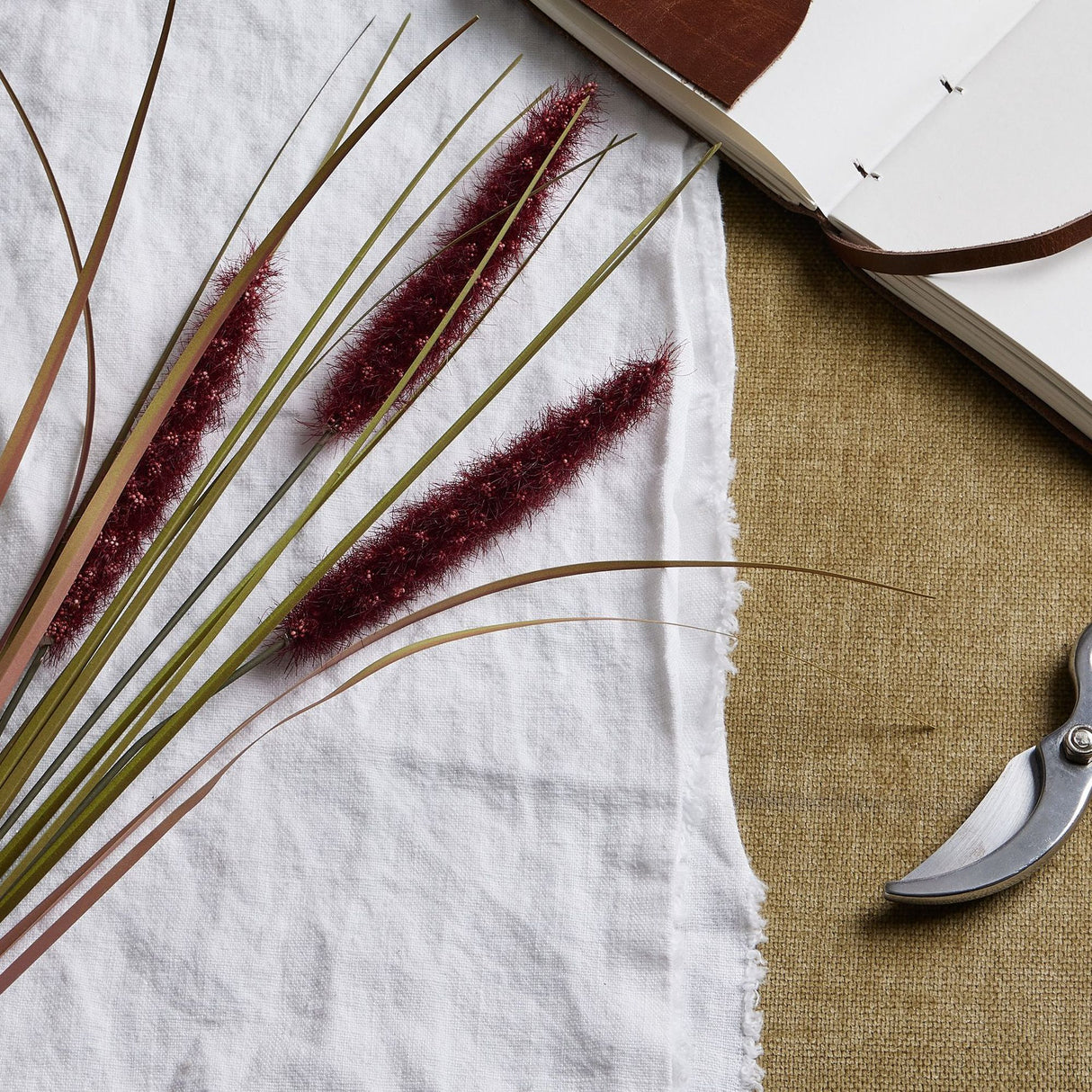Hill Interiors Ruby Triple Grass Stem with three red plumes on slender stems, on white linen beside an open leather journal and pruners.