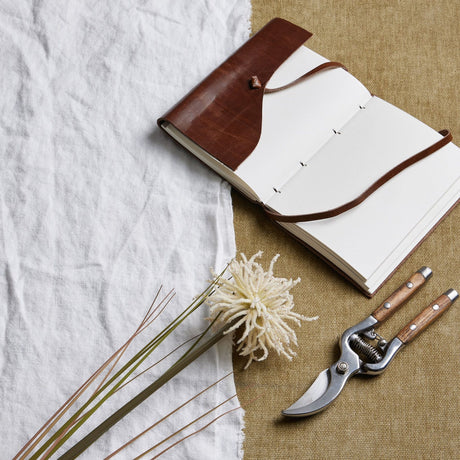 Hill Interiors Cream Aster Spider Chrysanthemum Stem styled on linen beside an open leather-bound journal and wooden-handled secateurs.