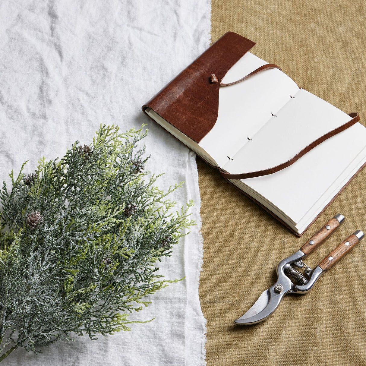 Hill Interiors Frosted Pine Spray Stem With Pinecones styled on white linen beside an open leather journal and wooden-handled secateurs.