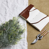 Hill Interiors Frosted Pine Spray Stem With Pinecones styled on white linen beside an open leather journal and wooden-handled secateurs.
