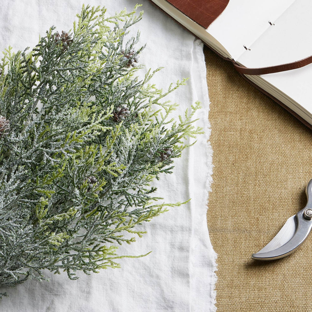 Hill Interiors Frosted Pine Spray Stem with pinecones laid on a white linen cloth beside an open leather-bound journal and metal secateurs.