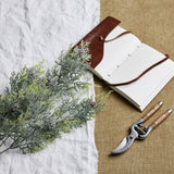 Hill Interiors Frosted Pine Tall Stem With Pinecones laid on white linen beside an open leather journal and wooden-handled secateurs on hessian.