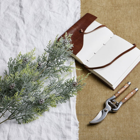 Hill Interiors Frosted Pine Tall Stem With Pinecones laid on white linen beside an open leather journal and wooden-handled secateurs on hessian.