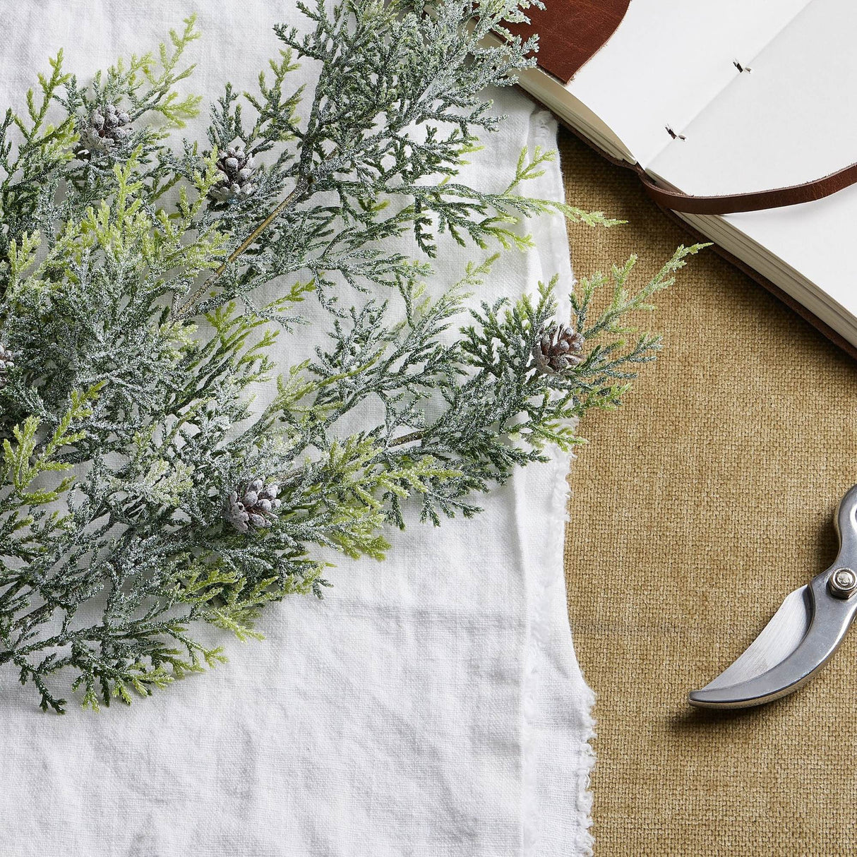 Hill Interiors Frosted Pine Tall Stem with pinecones laid on white linen beside an open leather journal and pruning shears on woven fabric.