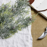 Hill Interiors Frosted Pine Tall Stem with pinecones laid on white linen beside an open leather journal and pruning shears on woven fabric.