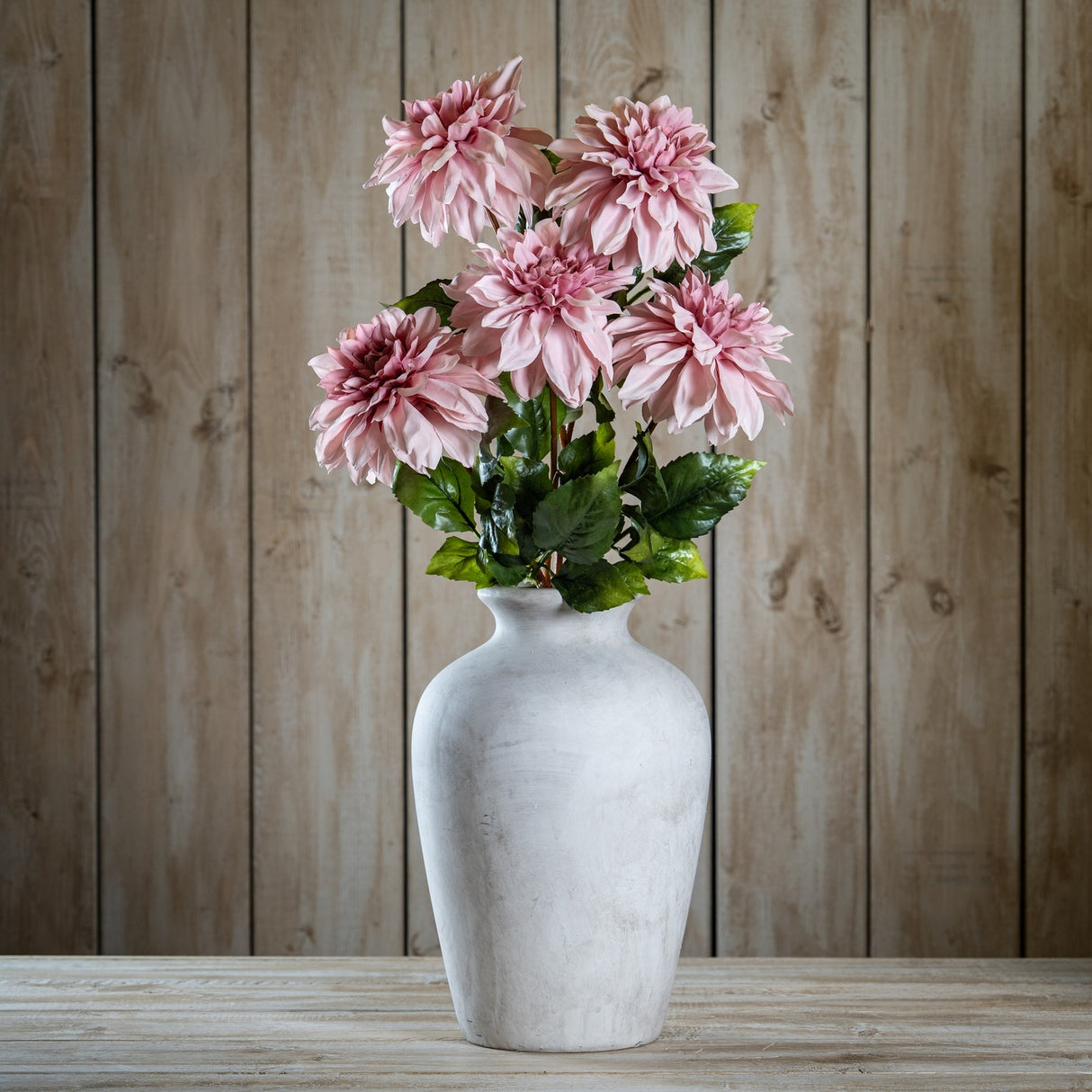 Hill Interiors Pink Dahlia faux flower stems with layered blush petals and green leaves arranged in a whitewashed stone vase against wood panelling.