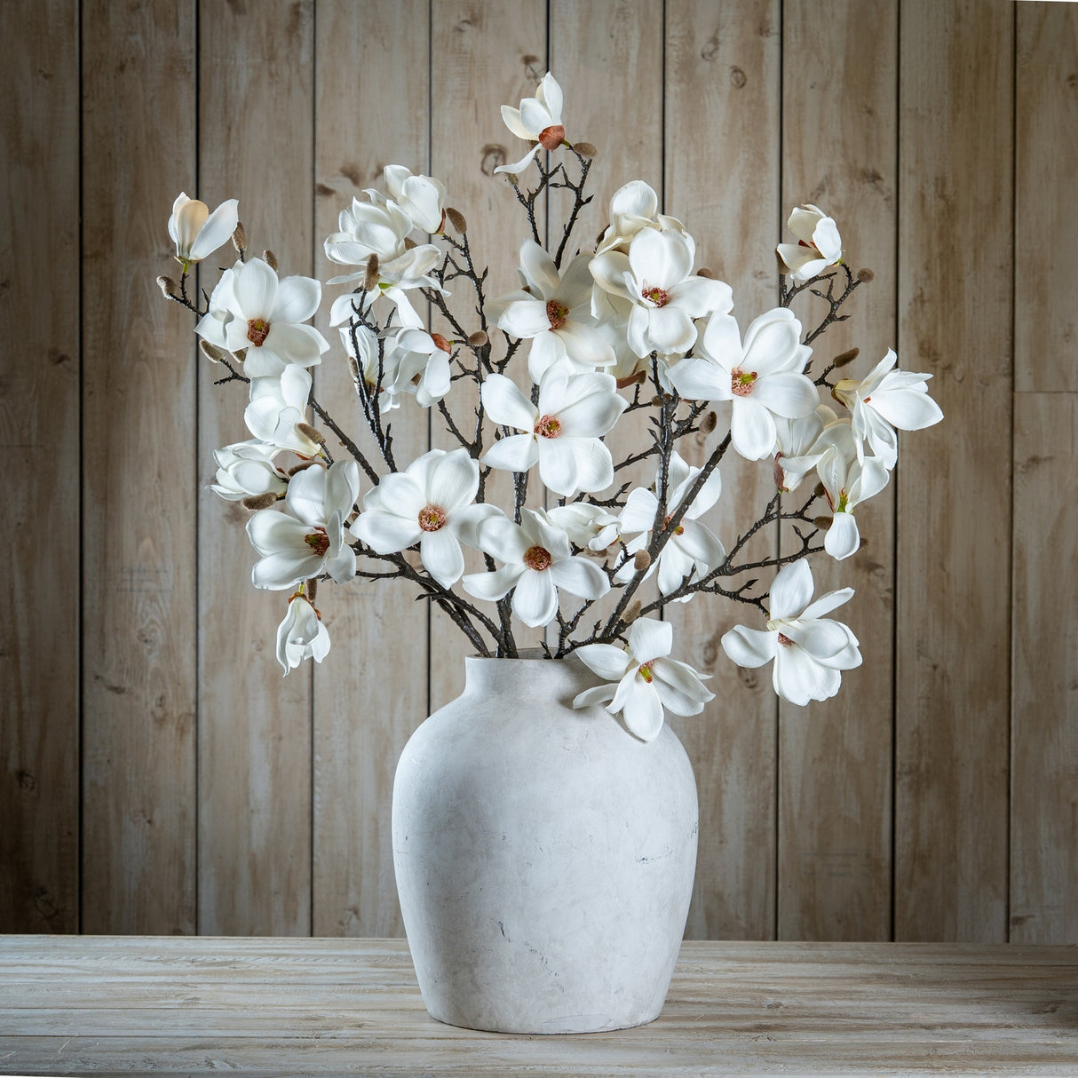 Hill Interiors White Magnolia Stems arranged in a stone ceramic vase against a rustic wood plank backdrop, showing layered petals and branching buds.