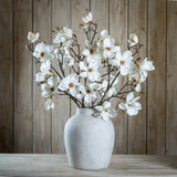 Hill Interiors White Magnolia Stems arranged in a stone ceramic vase against a rustic wood plank backdrop, showing layered petals and branching buds.