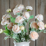 Hill Interiors Pink Ranunculus faux stems with layered blush petals and green foliage arranged in a white ceramic vase against a wooden plank backdrop.