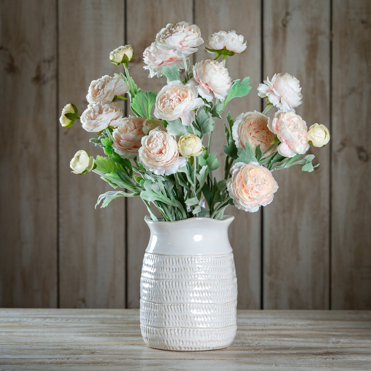 Hill Interiors Pink Ranunculus faux stems with layered blush petals and green foliage, arranged in a white textured ceramic vase against a wooden backdrop.