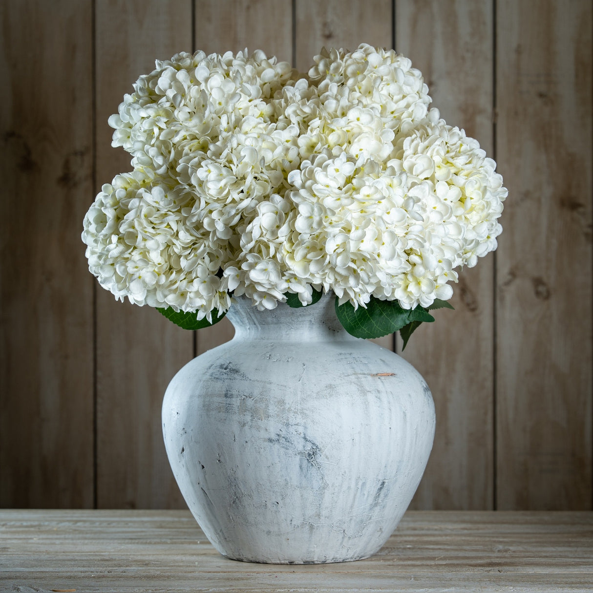 Hill Interiors White Allium faux stems arranged as rounded white hydrangea-like blooms in a distressed white stone vase against a wooden plank backdrop.