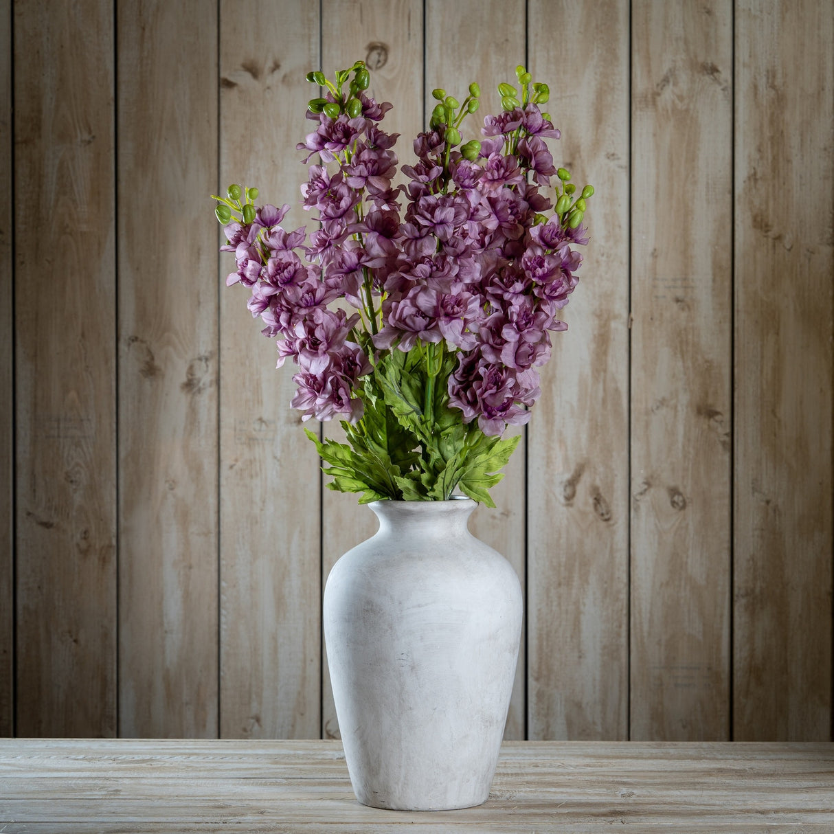 Hill Interiors Purple Elatum Delphinium faux stems with ruffled florets and green leaves arranged in a pale stone vase on wood.