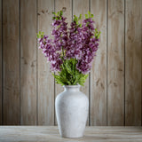 Hill Interiors Purple Elatum Delphinium faux stems with ruffled florets and green leaves arranged in a pale stone vase on wood.