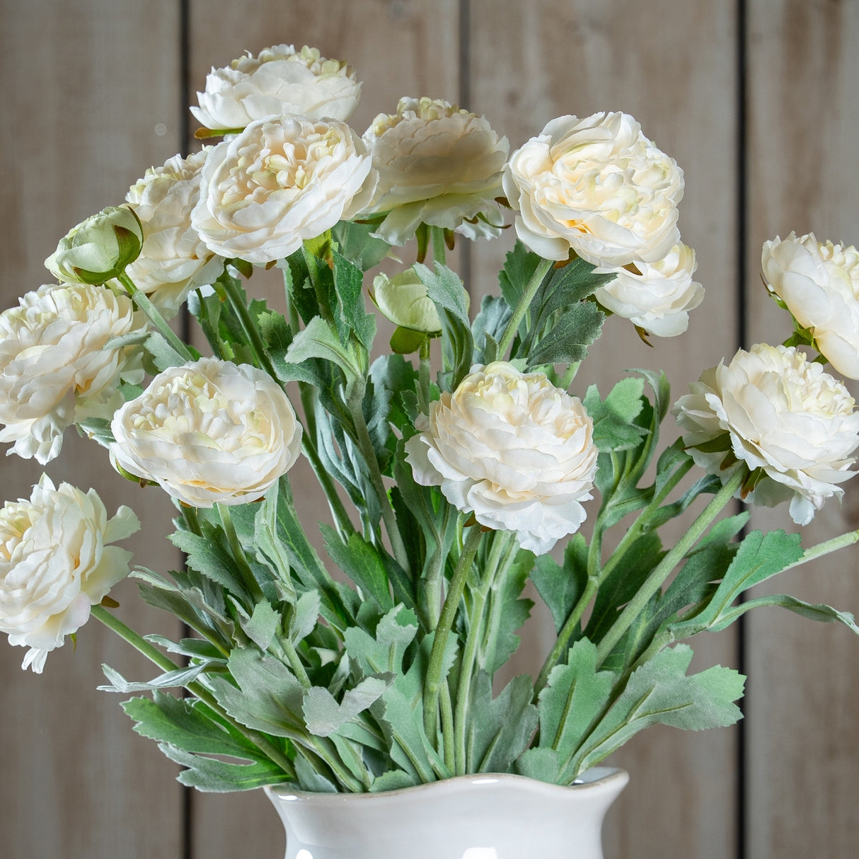 Hill Interiors White Ranunculus faux flower stems with ruffled cream-white blooms and green foliage, arranged in a white vase against a wood plank backdrop.