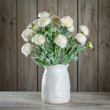 Hill Interiors White Ranunculus faux flower stems with ruffled cream blooms and green foliage arranged in a textured white ceramic jug on wood.
