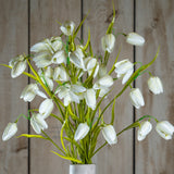 Hill Interiors White Fritillaria faux flower stem with nodding cream bell-shaped blooms and slim green foliage, displayed against a wooden plank background.