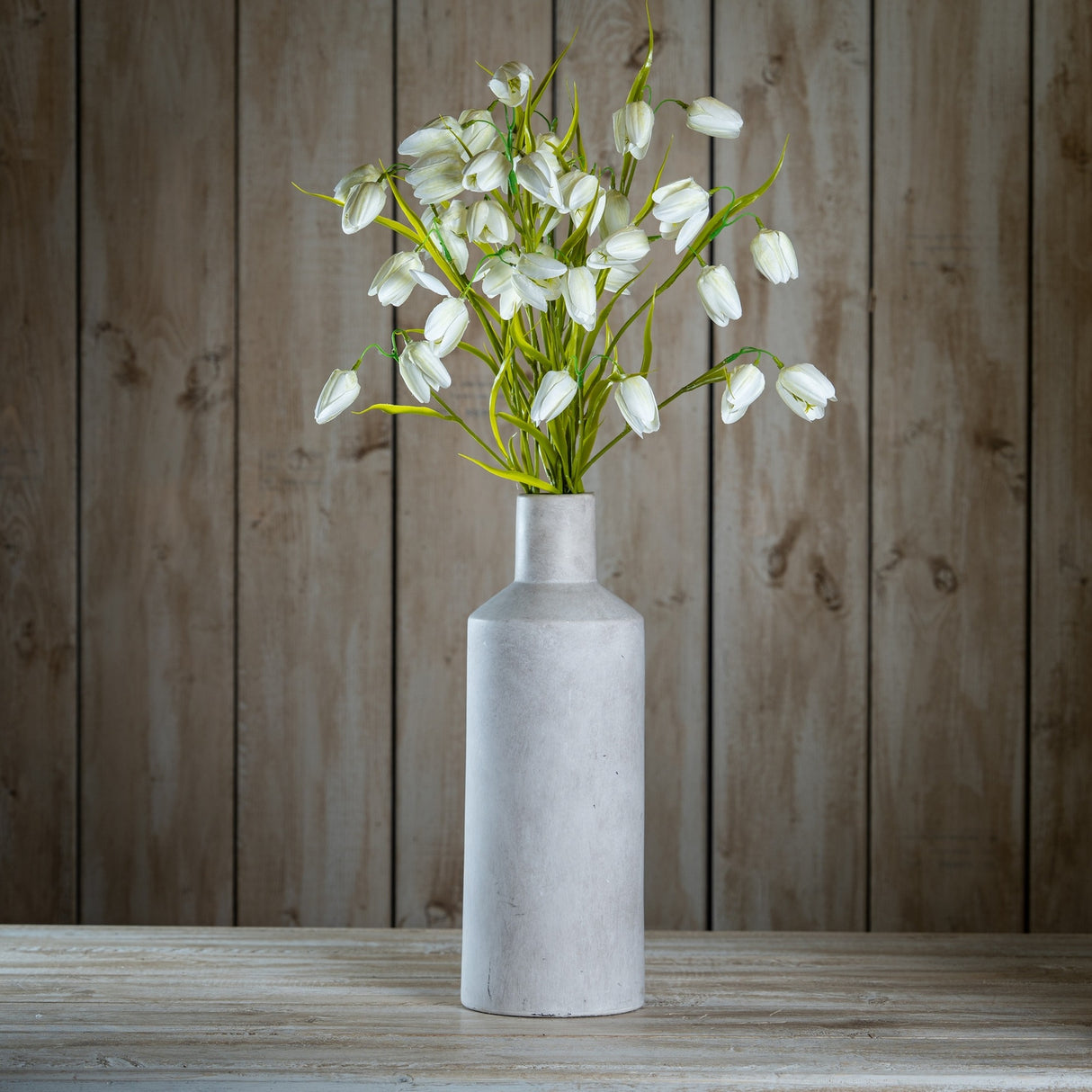 Hill Interiors White Fritillaria Stem with nodding bell-shaped blooms and slim green foliage arranged in a tall grey stone bottle vase against wood panelling.
