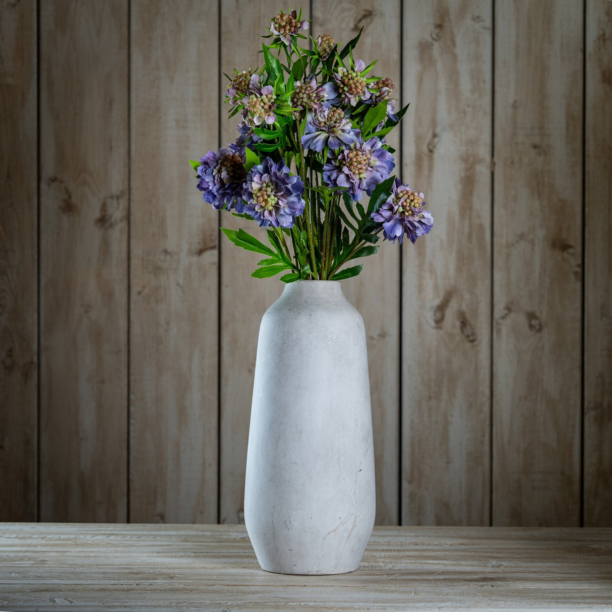 Hill Interiors Purple Scabious Stem arranged in a tall stone-effect vase on a wooden surface against a rustic timber plank background.
