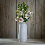 Hill Interiors Pale Pink Scabious Stems arranged in a tall stone vase on a wooden surface against a rustic timber plank backdrop.