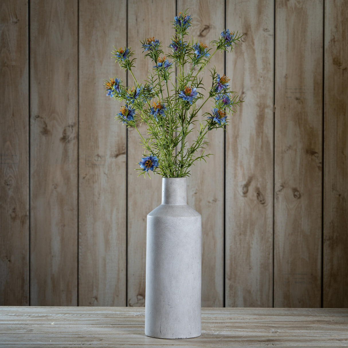 Hill Interiors Purple Nigella Love In A Mist faux stems with feathery bracts arranged in a tall grey stone bottle vase against wood panelling.