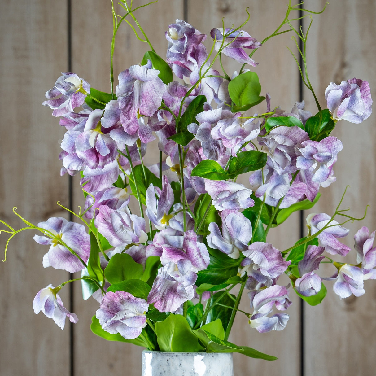 Purple Lilac Stem by Hill Interiors displayed in a speckled vase, with clustered lilac-toned florets and green leaves against a pale wood plank background.