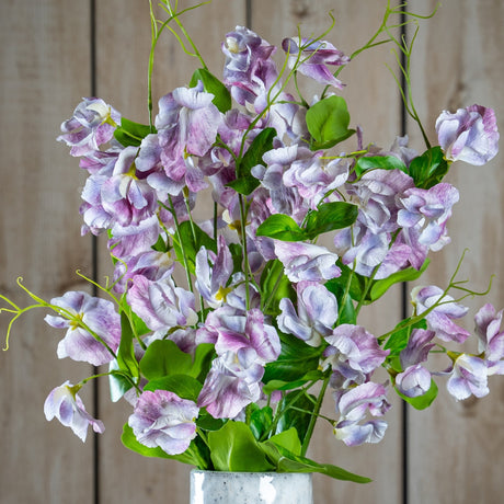 Purple Lilac Stem by Hill Interiors displayed in a speckled vase, with clustered lilac-toned florets and green leaves against a pale wood plank background.