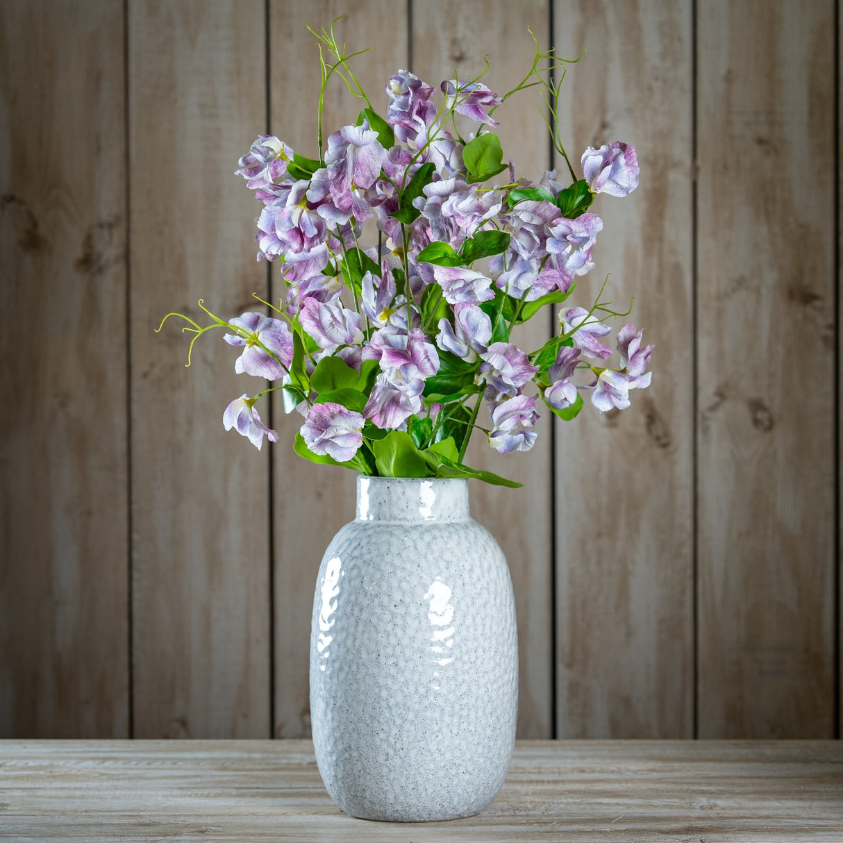 Hill Interiors Purple Lilac Stem arranged in a textured white ceramic vase with green leaves, set on a wooden surface against a plank wall.