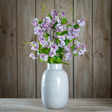 Hill Interiors Purple Lilac Stem arranged in a textured white ceramic vase with green leaves, set on a wooden surface against a plank wall.