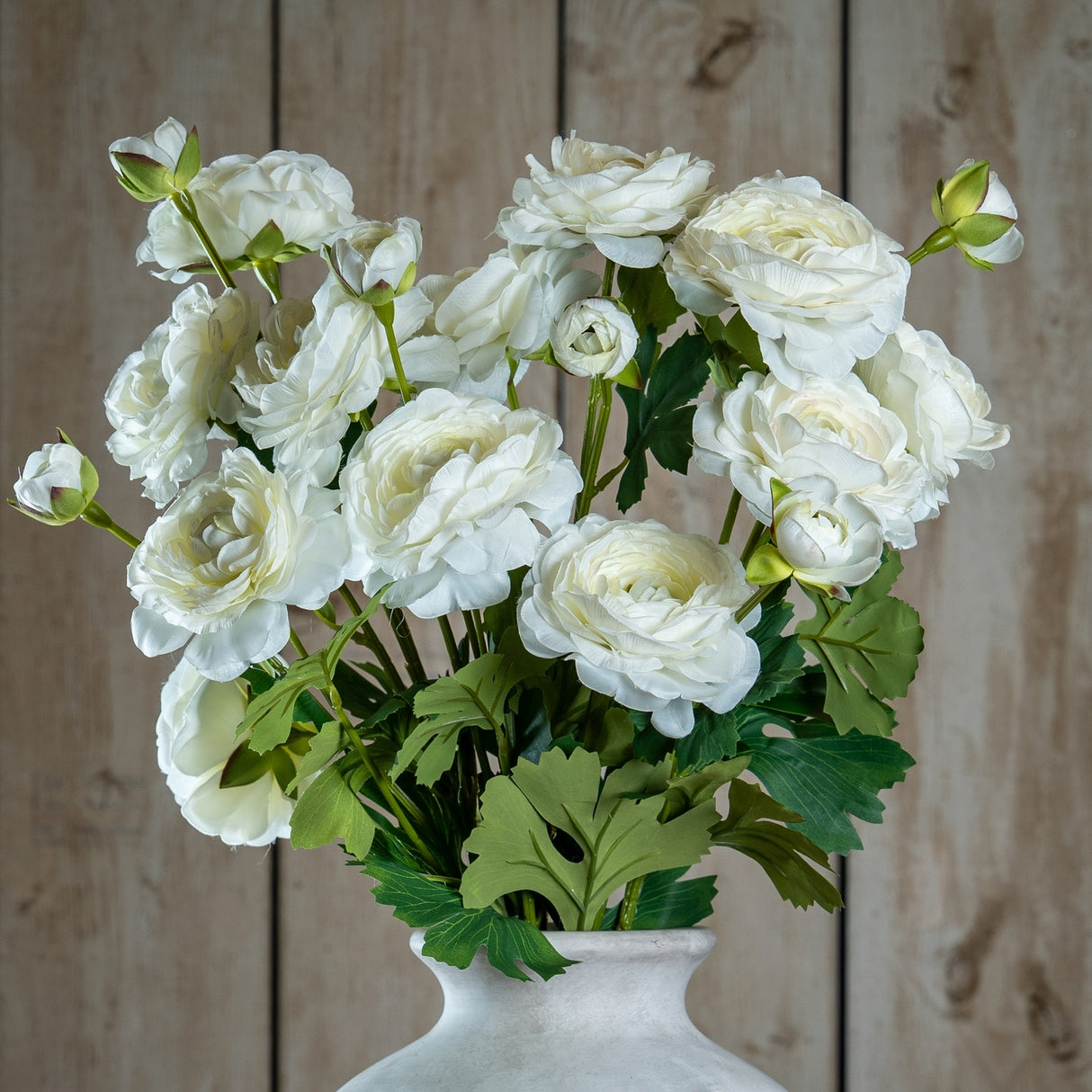 Hill Interiors Apricot Ranunculus faux stems arranged in a pale stone vase, showing layered white blooms and green foliage against a wood plank backdrop.