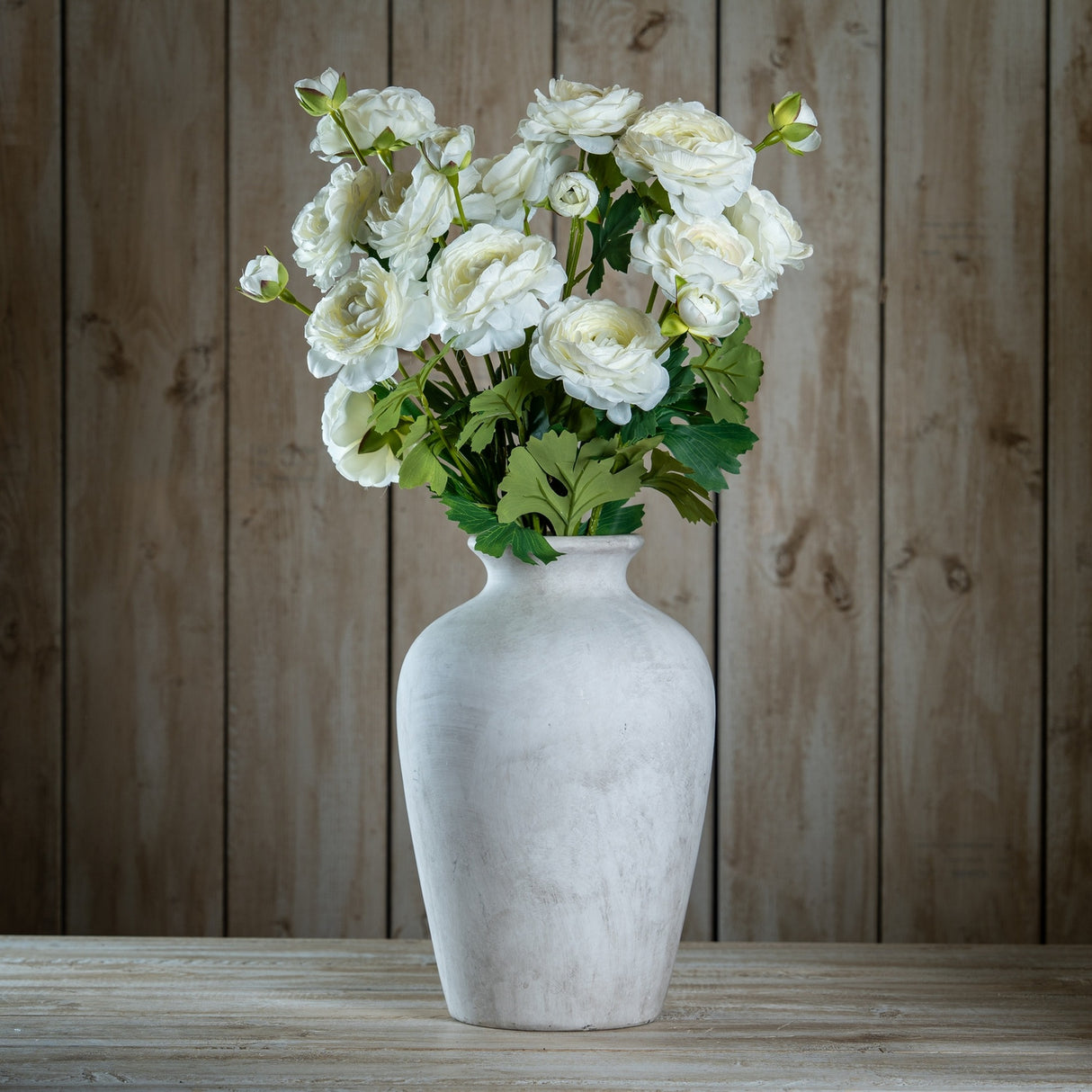 Hill Interiors Apricot Ranunculus faux stems arranged with green foliage in a whitewashed ceramic urn vase against a wooden plank backdrop.