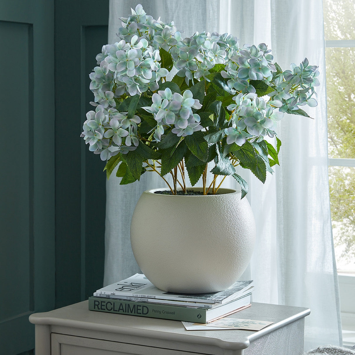 Hill Interiors faux hydrangea-style plant with pale blue-green blooms and lobed leaves in a round textured white pot, styled on books by a curtained window.
