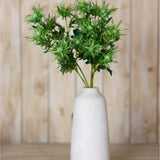 Hill Interiors Green Sea Holly Spray with spiky thistle-like heads and textured foliage arranged in a tall white ceramic vase against a wood panel backdrop.