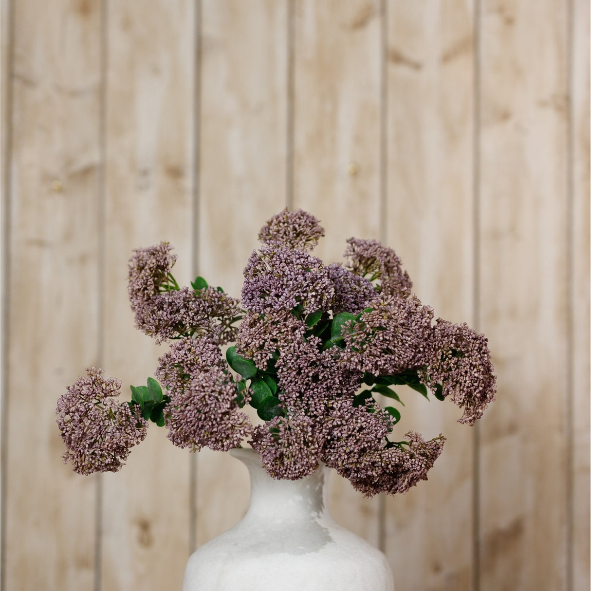 Hill Interiors Purple Sedum Spray with clustered mauve florets and green leaves arranged in a white ceramic vase against a light wood-panelled background.