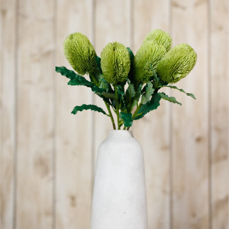 Hill Interiors Large Green Banksia Serrata faux stems arranged in a white ceramic vase against a pale wood panel background.
