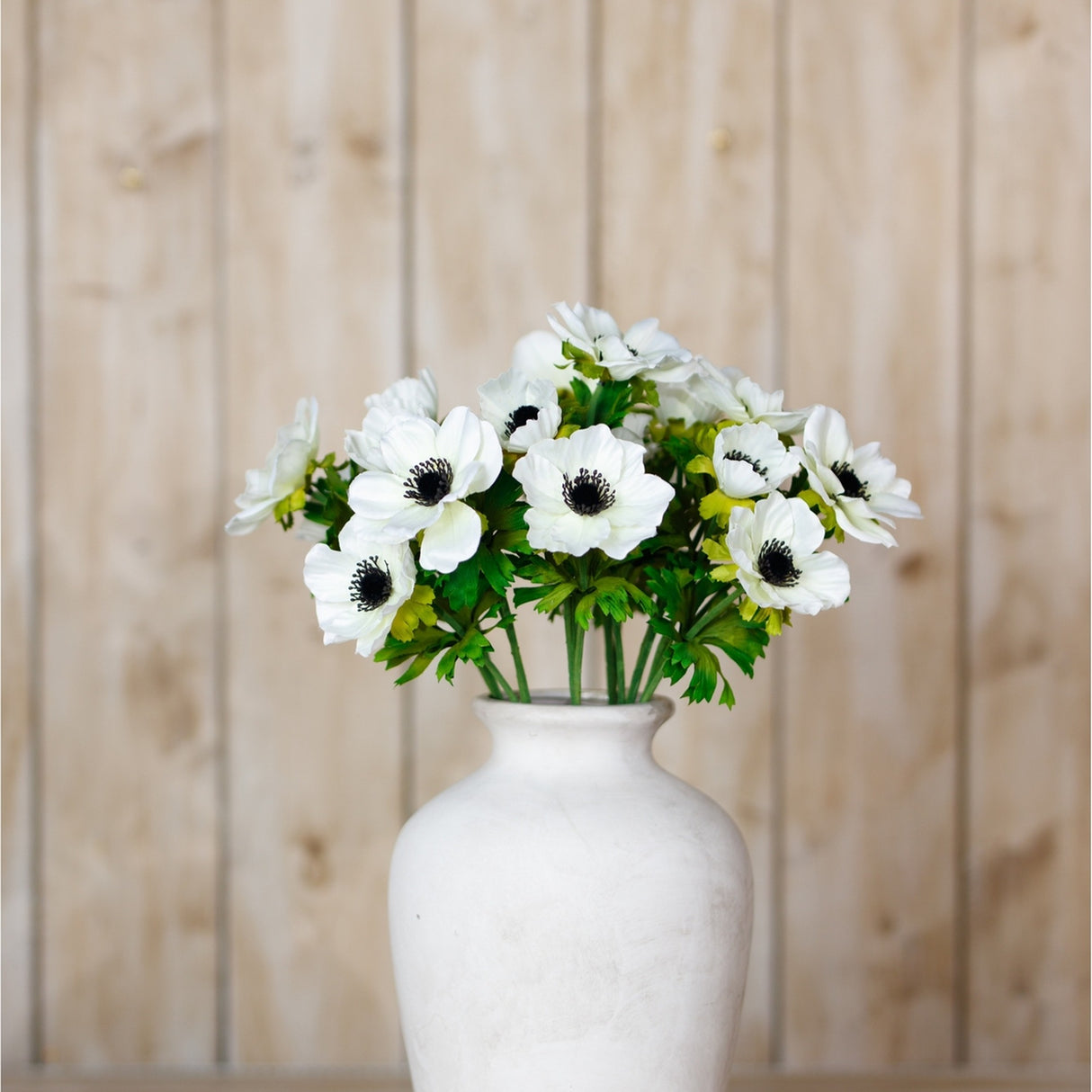 Hill Interiors White Anemone Spray artificial stems with dark centres arranged in a white ceramic vase against a light wood plank backdrop.