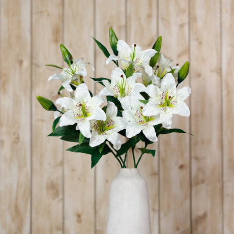 Hill Interiors White Madonna Lily Spray arranged in a tall white ceramic vase, showing open blooms and green buds against a light wood plank background.