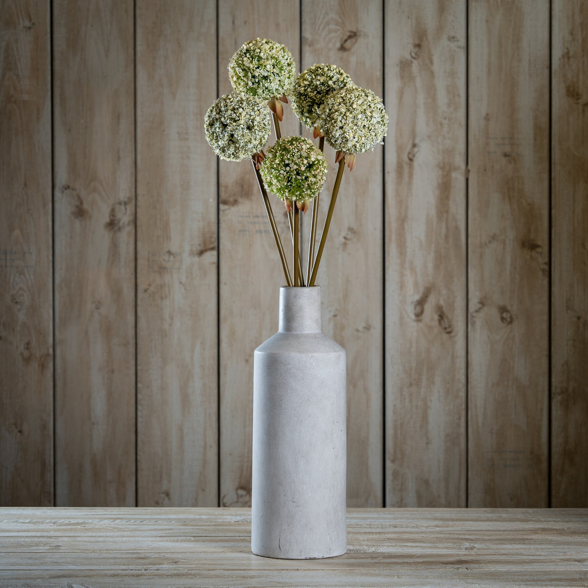 Hill Interiors Green Allium Stems arranged in a tall grey stoneware bottle vase on a wooden surface against a rustic timber plank background.