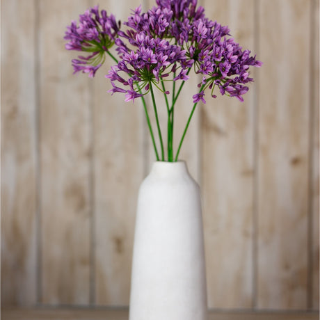 Hill Interiors Lilac Agapanthus Spray with clustered purple blooms on slim green stems, displayed in a tall white ceramic vase against wooden panelling.