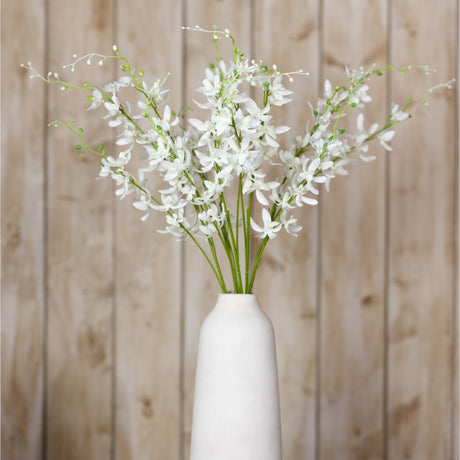 Hill Interiors White Cymbidium Spray arranged in a tall white ceramic vase, showing arching stems of small white orchid blooms against a wood plank backdrop.