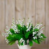 Hill Interiors Lily of the Valley Bunch faux plant with tiny white bell-shaped flowers and green leaves in a white vessel against wood panelling.