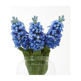Hill Interiors Blue Hyacinth Stem trio of faux plastic flower spikes with clustered blue florets and green leaves in a clear glass vase on a white background.