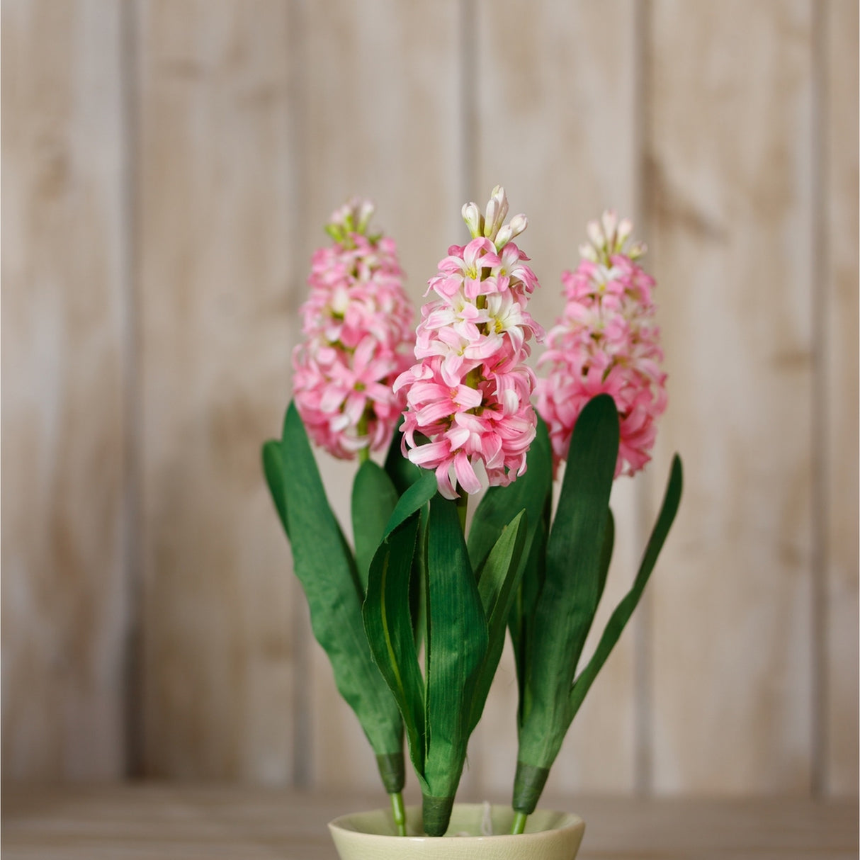 Hill Interiors Pink Hyacinth Stem faux plant with three clustered pink blooms and green leaves in a pale pot against a light wood panel background.