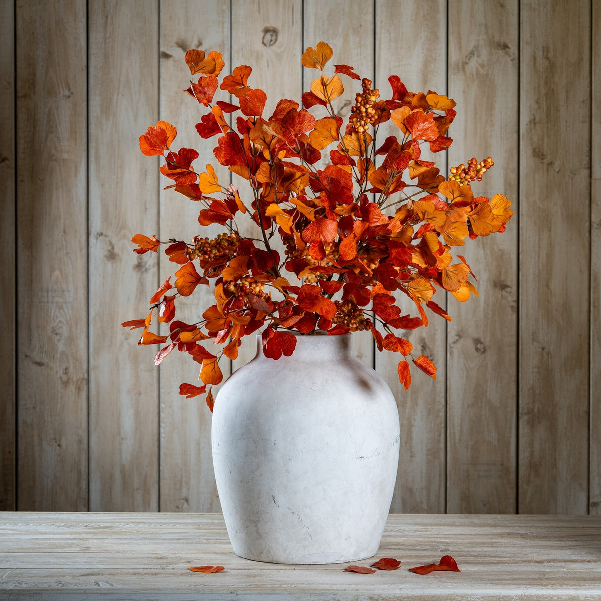 Hill Interiors Large Autumn Spray With Berries arranged in a stone vase, showing layered orange and brown foliage against a wooden plank backdrop.