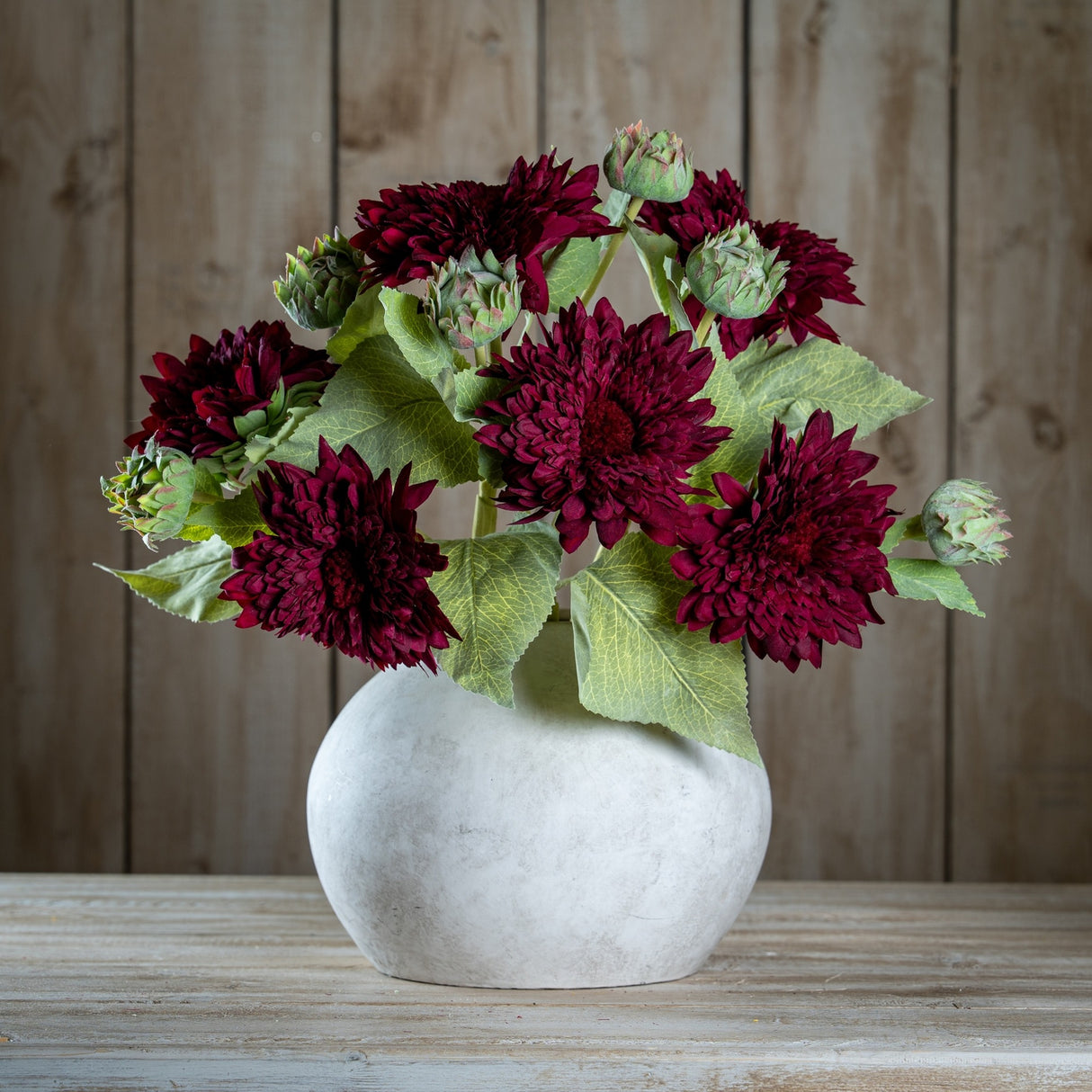 Hill Interiors Dahlia Burgundy Raven Stems arranged with green buds and leaves in a rounded matte stone vase on a wooden surface against timber panels.