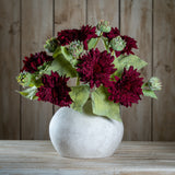 Hill Interiors Dahlia Burgundy Raven Stems arranged with green buds and leaves in a rounded matte stone vase on a wooden surface against timber panels.