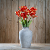 Hill Interiors Flame Orange Parrot Tulips with ruffled red and yellow petals arranged in a grey stone vase on a wooden surface against a plank wall.
