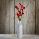 Hill Interiors Fireball Calla Lily faux stems with deep red trumpet blooms displayed in a tall grey ceramic vase against a wooden plank backdrop.
